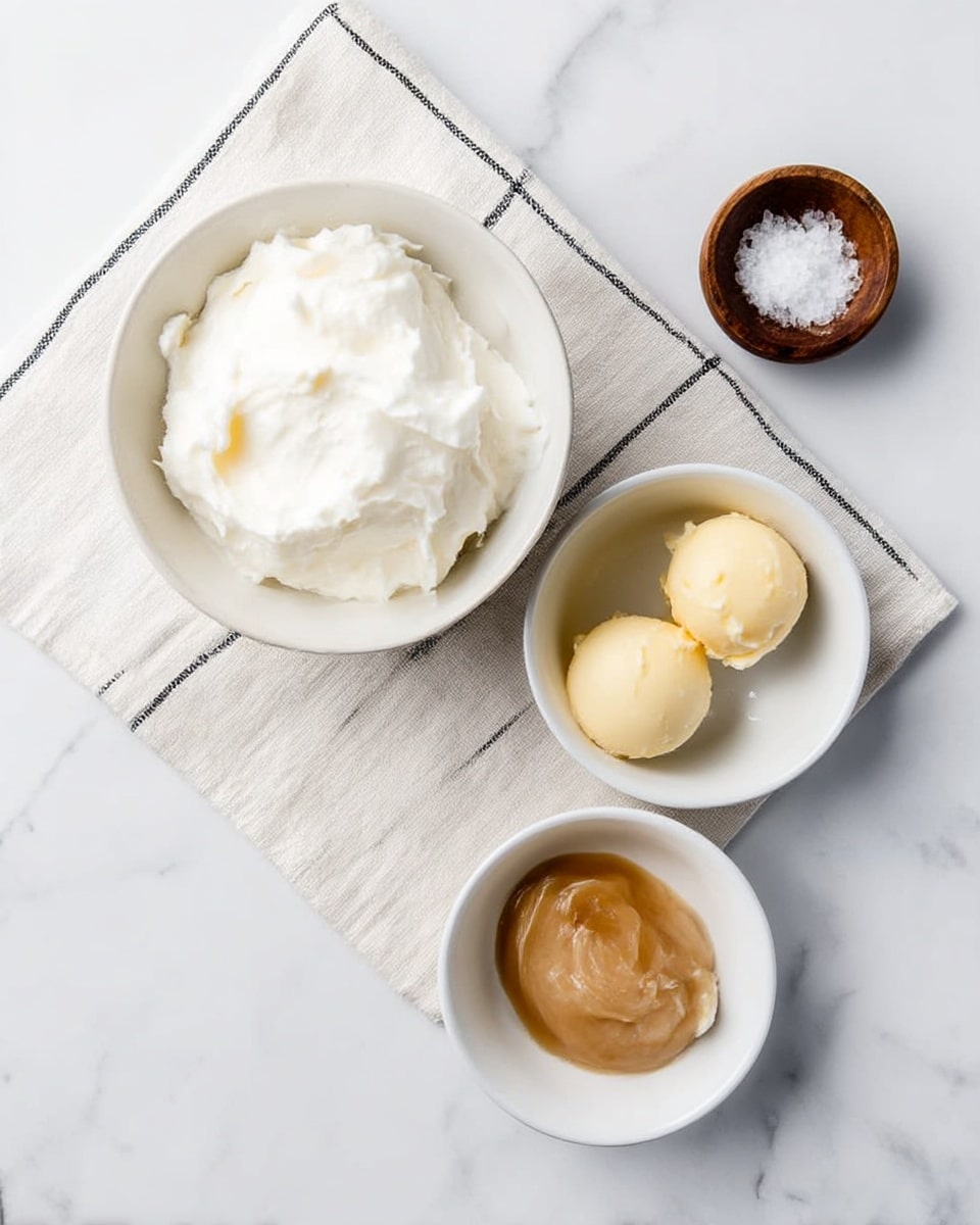 The image shows three white bowls on a white marbled surface with a white cloth that has black lines underneath them. The largest bowl contains a thick, white creamy substance with a soft and smooth texture. To the right, a smaller white bowl holds two small, round dollops of pale yellow butter with a smooth surface. Below these is the smallest white bowl containing a light brown, thick sauce spread flat and shiny. A small wooden bowl with coarse salt is also placed near the top right corner of the cloth. photo taken with an iphone --ar 4:5 --v 7