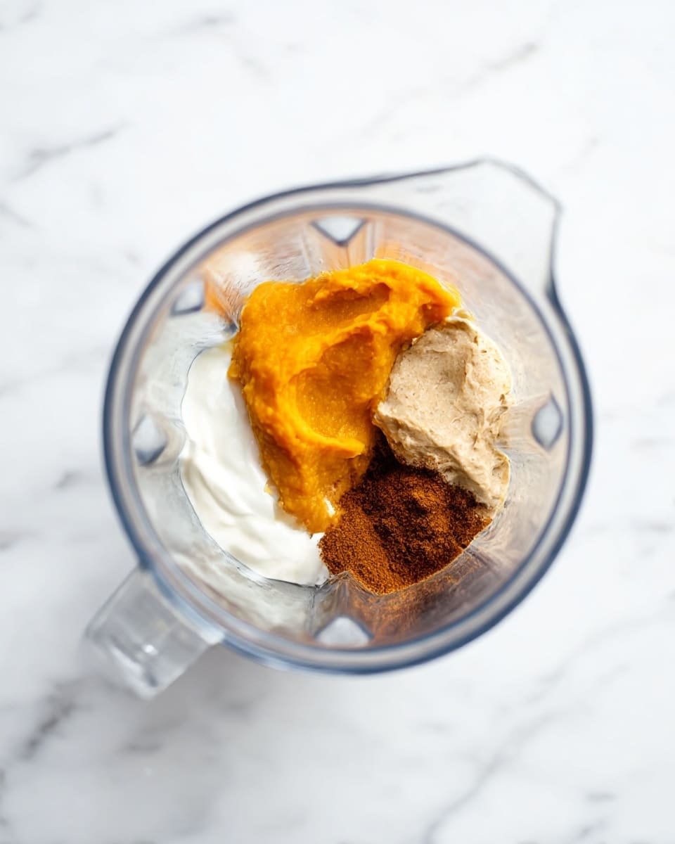 The image shows a clear blender jar from above, sitting on a white marbled surface. Inside the blender, there are five separate ingredients arranged in sections: bright orange smooth pumpkin puree on the left, a dollop of creamy white yogurt at the bottom, a light beige grainy paste at the right, a brown nut butter with a slightly rough texture toward the upper right, and a pile of dark brown ground spice in the center. The blender is clean and transparent, clearly showing each ingredient's color and texture. The photo taken with an iphone --ar 4:5 --v 7