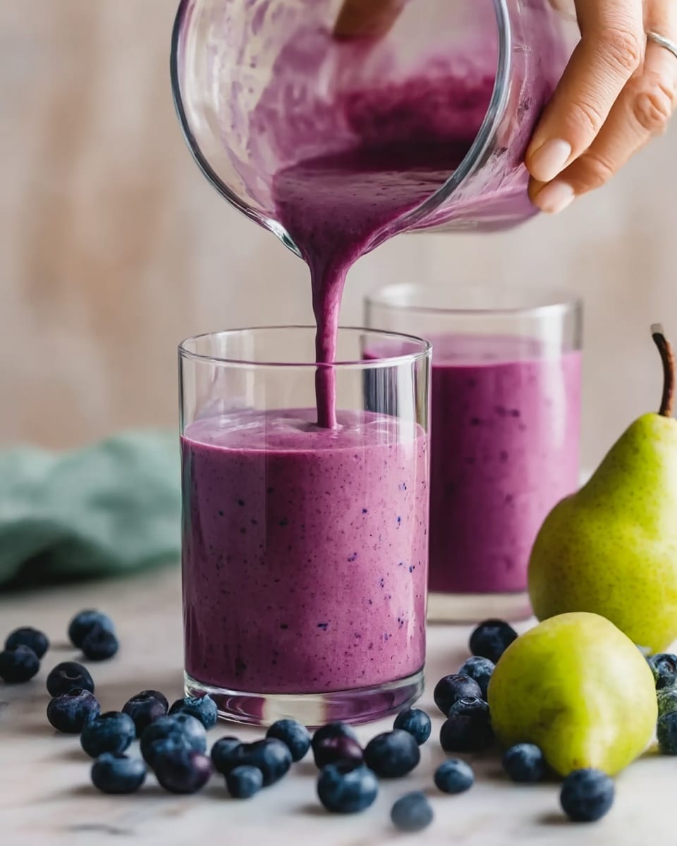 A glass filled with a thick purple smoothie is in the front, with a second glass filled with the same smoothie slightly behind it. A woman's hand is pouring more of the smooth purple smoothie into the glass in the front. Both glasses are clear and sit on a white marbled surface scattered with fresh blueberries and a green pear to the right side. The smoothie has a smooth texture with tiny specks visible inside. Photo taken with an iphone --ar 4:5 --v 7