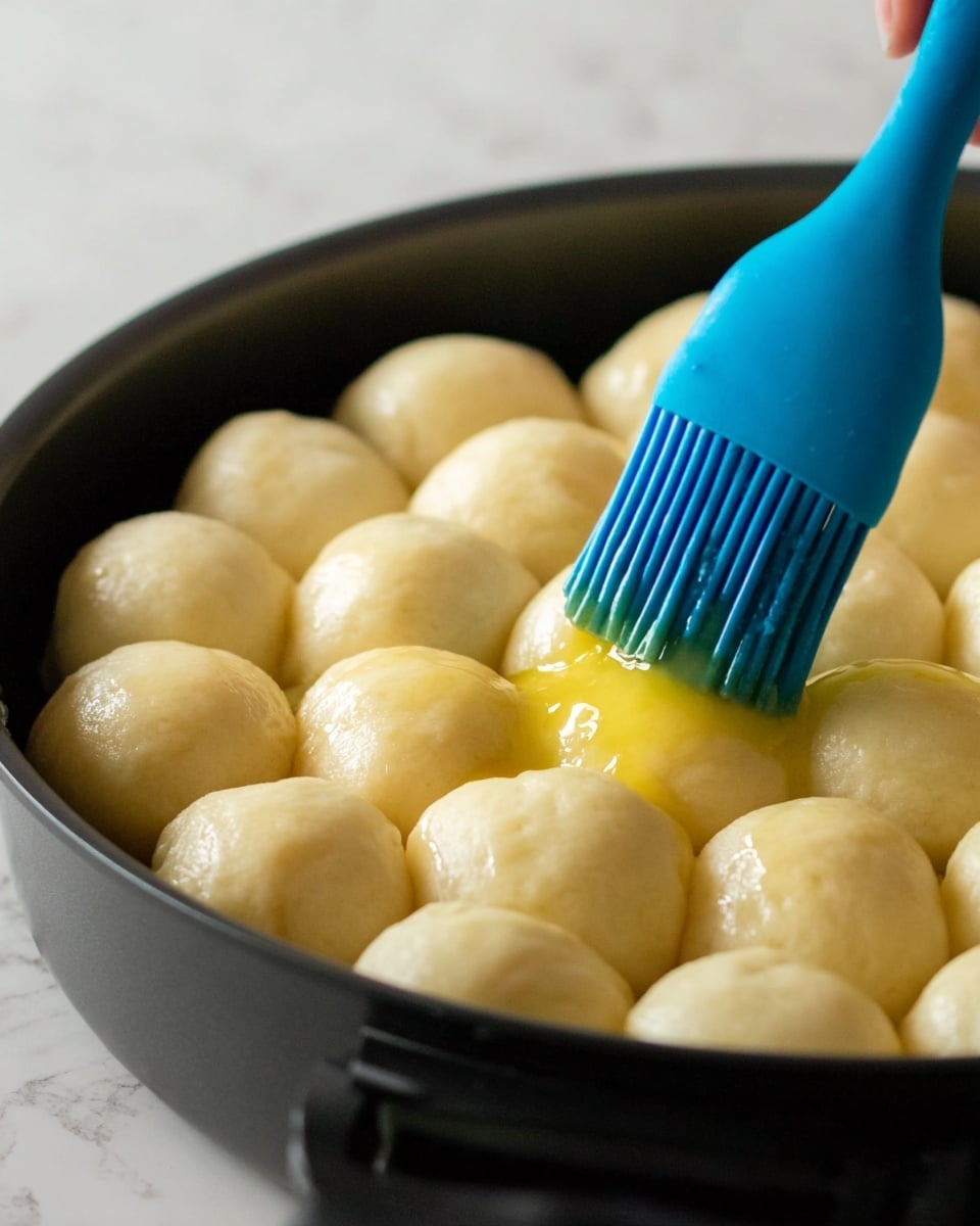 A close-up image shows a round black pan filled with small, smooth dough balls arranged closely together in one layer. A woman’s hand is holding a blue silicone brush that is spreading a shiny yellow liquid, likely butter or egg wash, over the top of the pale dough balls, making them look moist and glossy. The background is softly blurred with a white marbled texture visible beneath the pan. Photo taken with an iphone --ar 4:5 --v 7