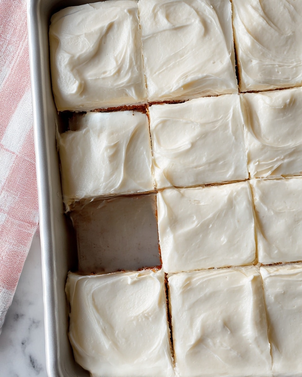 The image shows a close-up top view of a rectangular white tray with nine square pieces of cake inside. Each piece is covered with a thick, smooth layer of white frosting that has a creamy texture with soft swirls and slight peaks. The cake pieces are separated by straight cuts that reveal a thin, light brown edge beneath the frosting. The tray rests on a white marbled surface with a light pink and white striped cloth partially visible on the left side. photo taken with an iphone --ar 4:5 --v 7