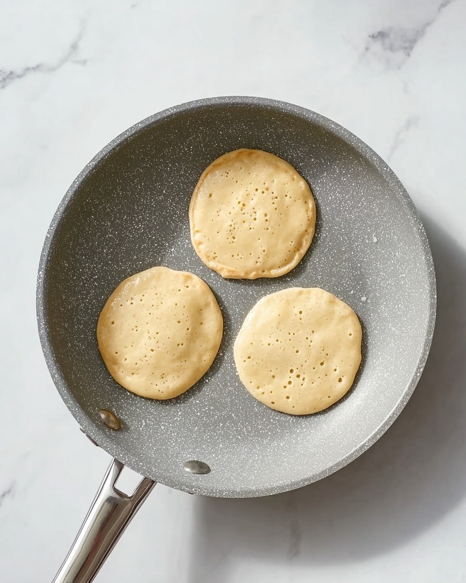 A grey speckled frying pan holds three small, round pancake batter circles cooking on its surface, each pancake showing bubbles as they start to cook and edges that are slightly uneven. The pan is seen from above, placed on a white marbled surface. The handle of the pan is thin and metallic, extending out to the left side of the image. photo taken with an iphone --ar 4:5 --v 7