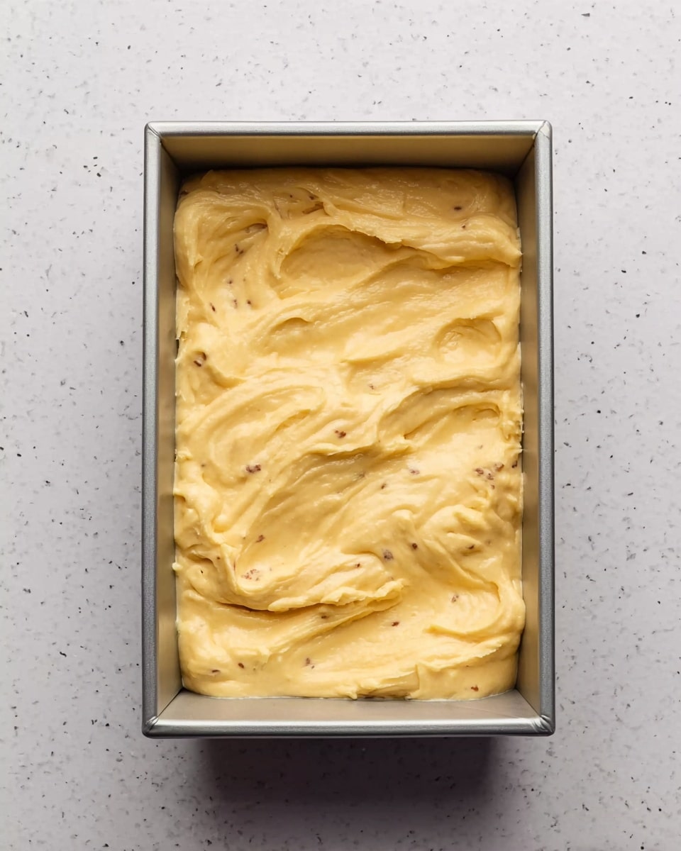 A smooth, thick, yellow batter with small brown spots is spread evenly inside a silver rectangular baking pan, filling it about halfway. The surface of the batter is somewhat uneven with gentle swirls and slight mounds visible. The pan sits on a white marbled texture. photo taken with an iphone --ar 4:5 --v 7