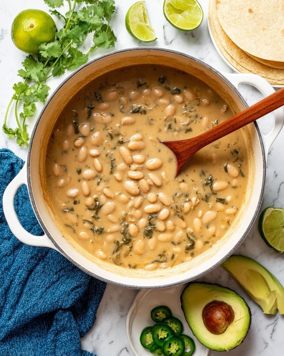 A white pot filled with a thick creamy beige bean soup with visible whole white beans and bits of greens, the texture looks smooth with some chunkiness. A wooden spoon is resting inside the soup, slightly stirring it. Around the pot, on a white marbled surface, there are halves of green lime, fresh cilantro sprigs, two avocado halves with dark brown seeds, a small white plate holding sliced green jalapeños, and white tortillas stacked in the corner. A folded blue cloth is placed near the pot. photo taken with an iphone --ar 4:5 --v 7