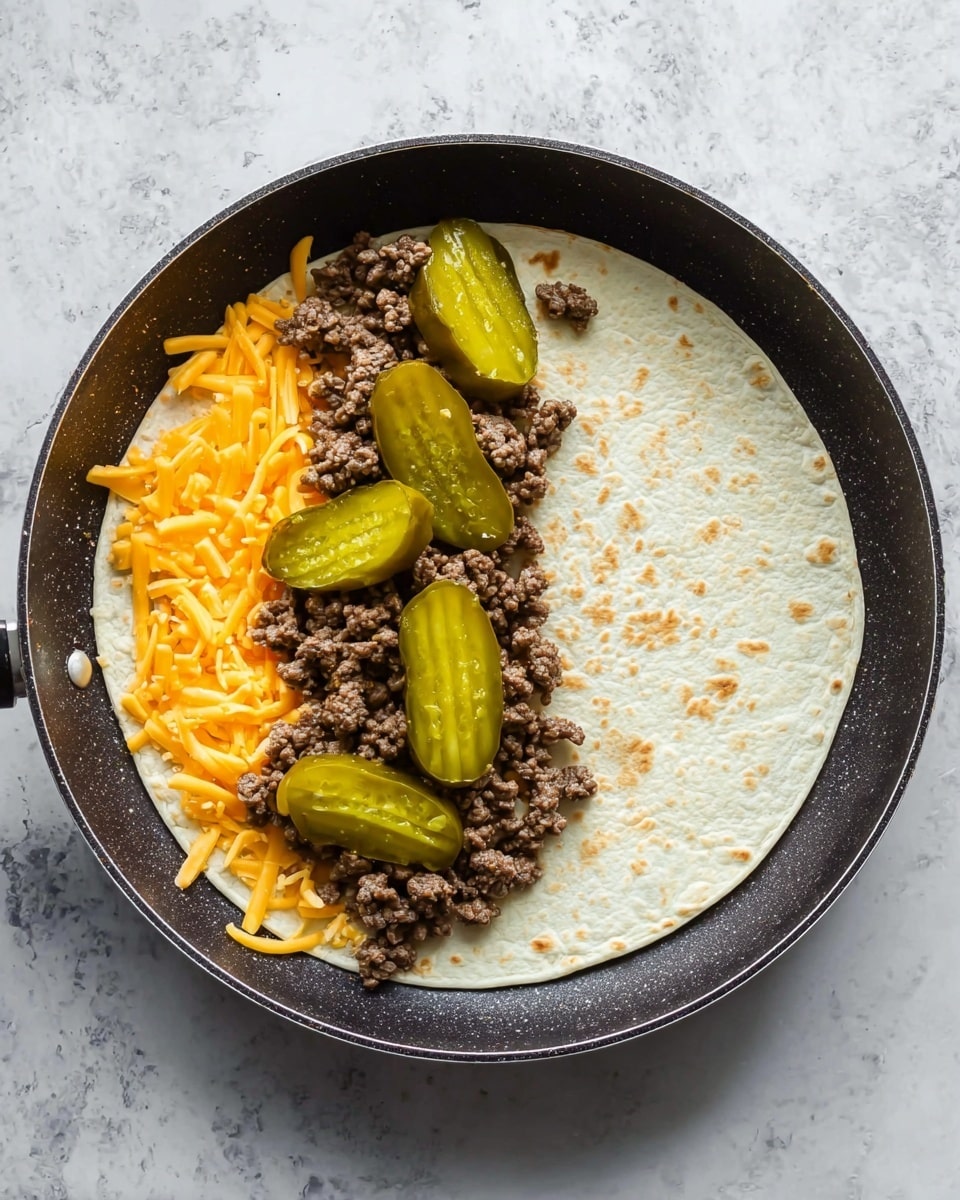 A single white tortilla lies flat inside a dark frying pan on a white marbled surface. On the left half of the tortilla, there is a layer of shredded orange cheddar cheese spread evenly. On top of the cheese sits a layer of cooked ground beef, brown in color and crumbly in texture. Five crinkled, bright green pickle slices are placed on top of the beef layer. The right half of the tortilla is empty, showing its smooth, slightly textured surface. Photo taken with an iphone --ar 4:5 --v 7