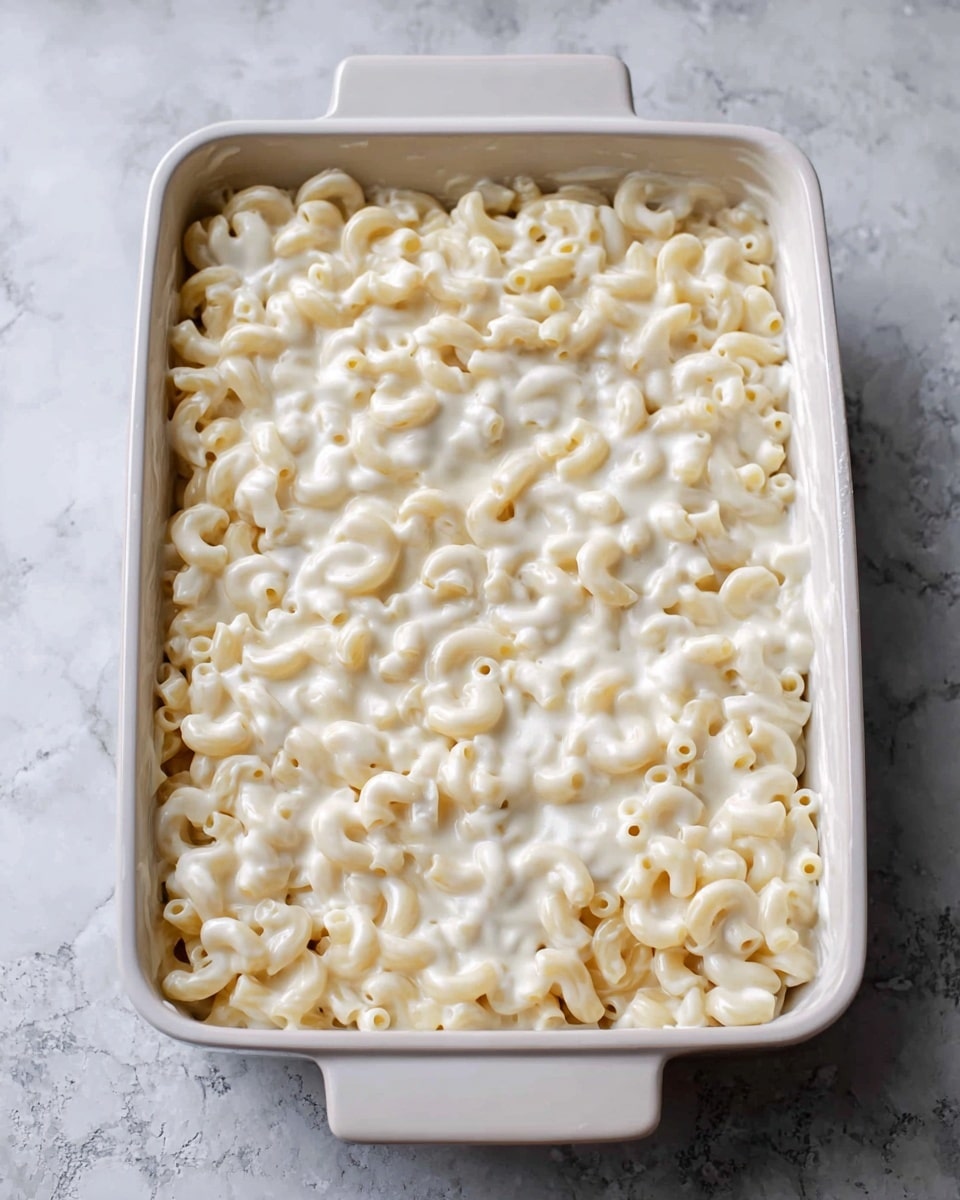 A white rectangular baking dish filled with creamy pasta inside, showing one thick layer of elbow macaroni covered in smooth white sauce. The shapes of the macaroni are soft and slightly shiny, all evenly coated with the sauce that has a light, creamy texture. The dish is placed on a white marbled surface. photo taken with an iphone --ar 4:5 --v 7