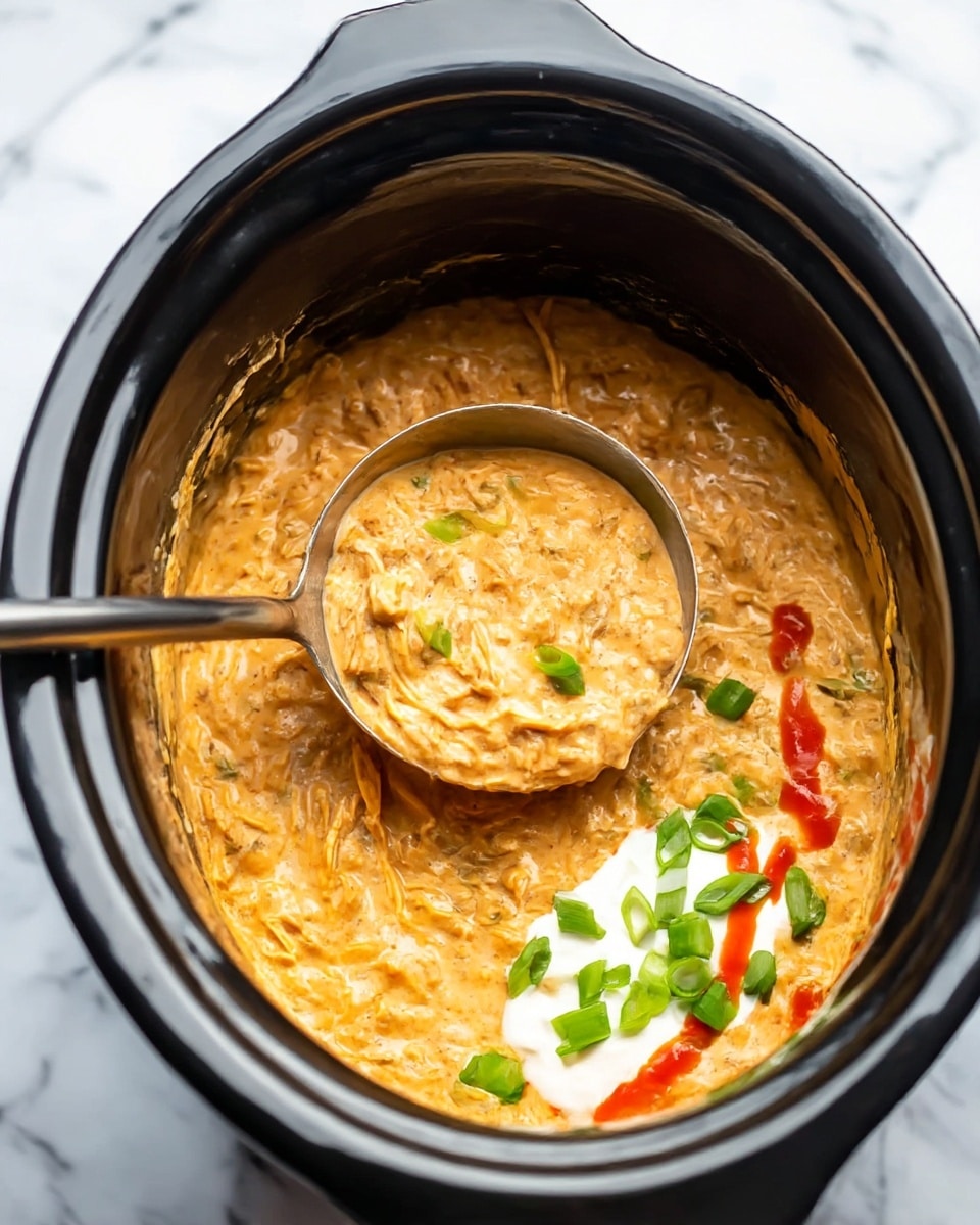 The image shows a black slow cooker filled with a creamy, orange-colored dip with a thick and chunky texture. Inside the dip, pieces of shredded meat are visible, mixed evenly throughout the creamy base. A large shiny metal ladle rests inside the slow cooker, scooping up some of the dip. On the right side image, the dip is topped with small green slices of chopped green onions, a dollop of white sour cream near the ladle’s edge, and a drizzle of bright red hot sauce, adding color contrast. The background is a white marbled surface. photo taken with an iphone --ar 4:5 --v 7