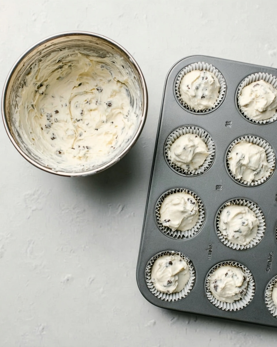 The image shows two parts: on the left, a silver metal bowl with white cream mixed with small black pieces inside, placed on a white marbled surface. On the right, a grey metal muffin tray holding twelve white paper liners, each filled with a portion of the same white cream mixture, also set on the white marbled surface. photo taken with an iphone --ar 4:5 --v 7