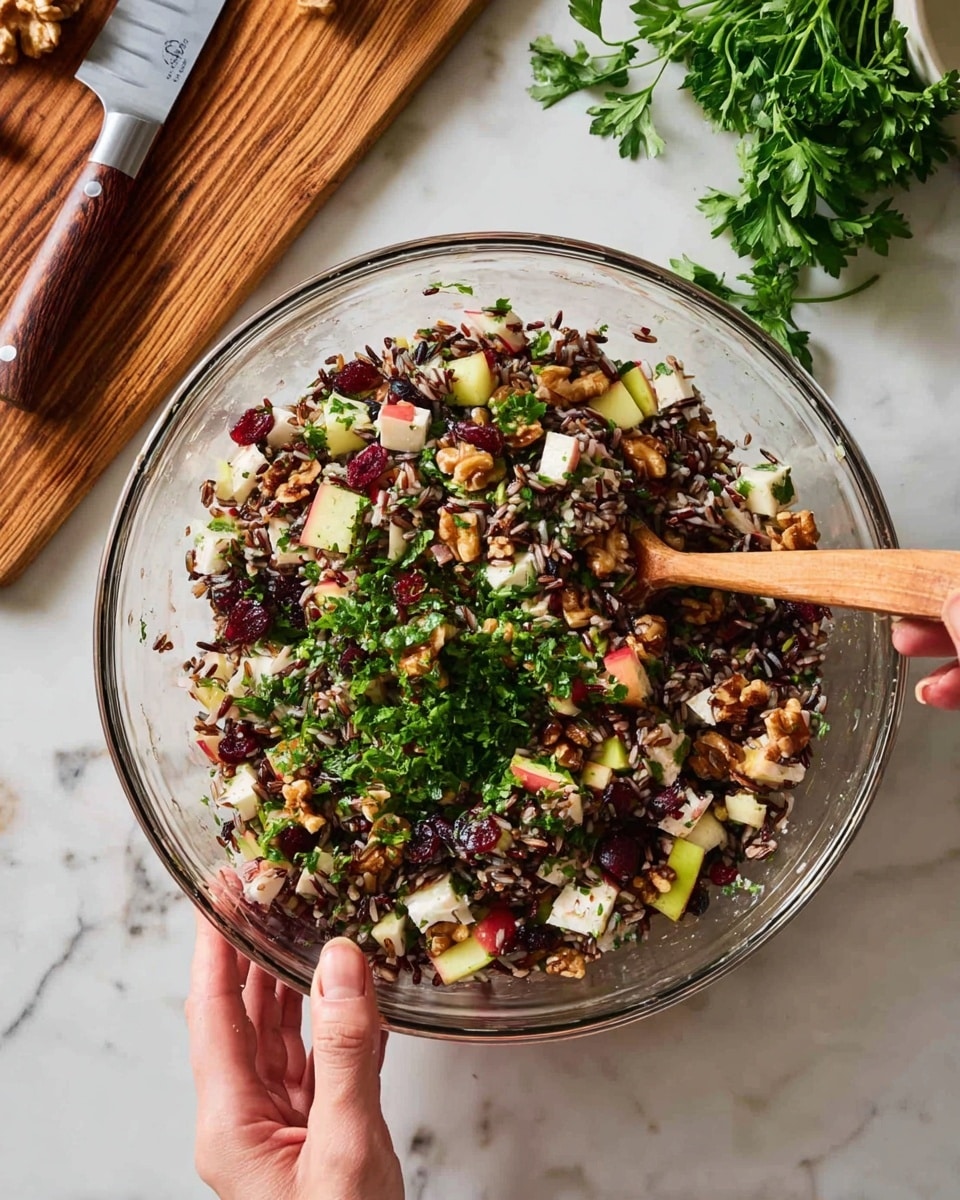 A clear glass bowl filled with a mixed salad that has three main layers: the bottom layer is made of dark and wild rice grains, mixed with small white pieces of soft cheese; the middle layer has diced red apple chunks, dark dried cranberries, and light brown nuts; the top layer is fresh, bright green chopped herbs spread across the salad. A wooden spoon with a light brown handle is inside the bowl, partly covered with the mixture, and a woman's hand is holding the bowl from the side. The bowl is placed on a white marbled surface, with a wooden cutting board, fresh green parsley, and a chef's knife with a wooden handle visible near the top. Photo taken with an iphone --ar 4:5 --v 7