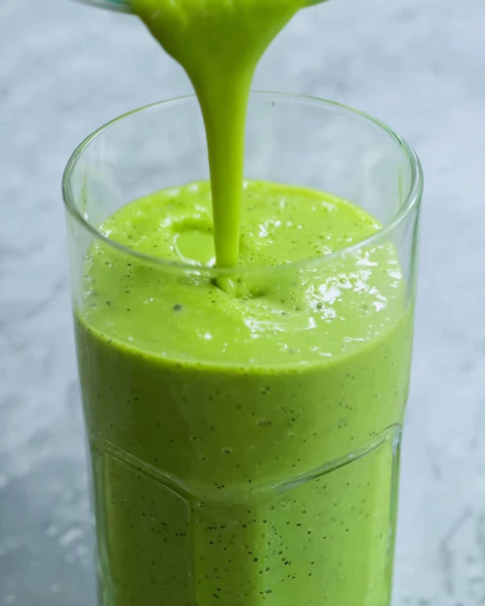 The image shows a clear glass filled almost to the top with a thick, bright green smoothie that has tiny dark green specks throughout the mixture. The smoothie is being poured into the glass from above, forming a smooth, flowing stream that creates a slight swirl and bubbles on the surface inside the glass. The glass is simple and transparent, set against a background with a white marbled texture. Photo taken with an iphone --ar 4:5 --v 7