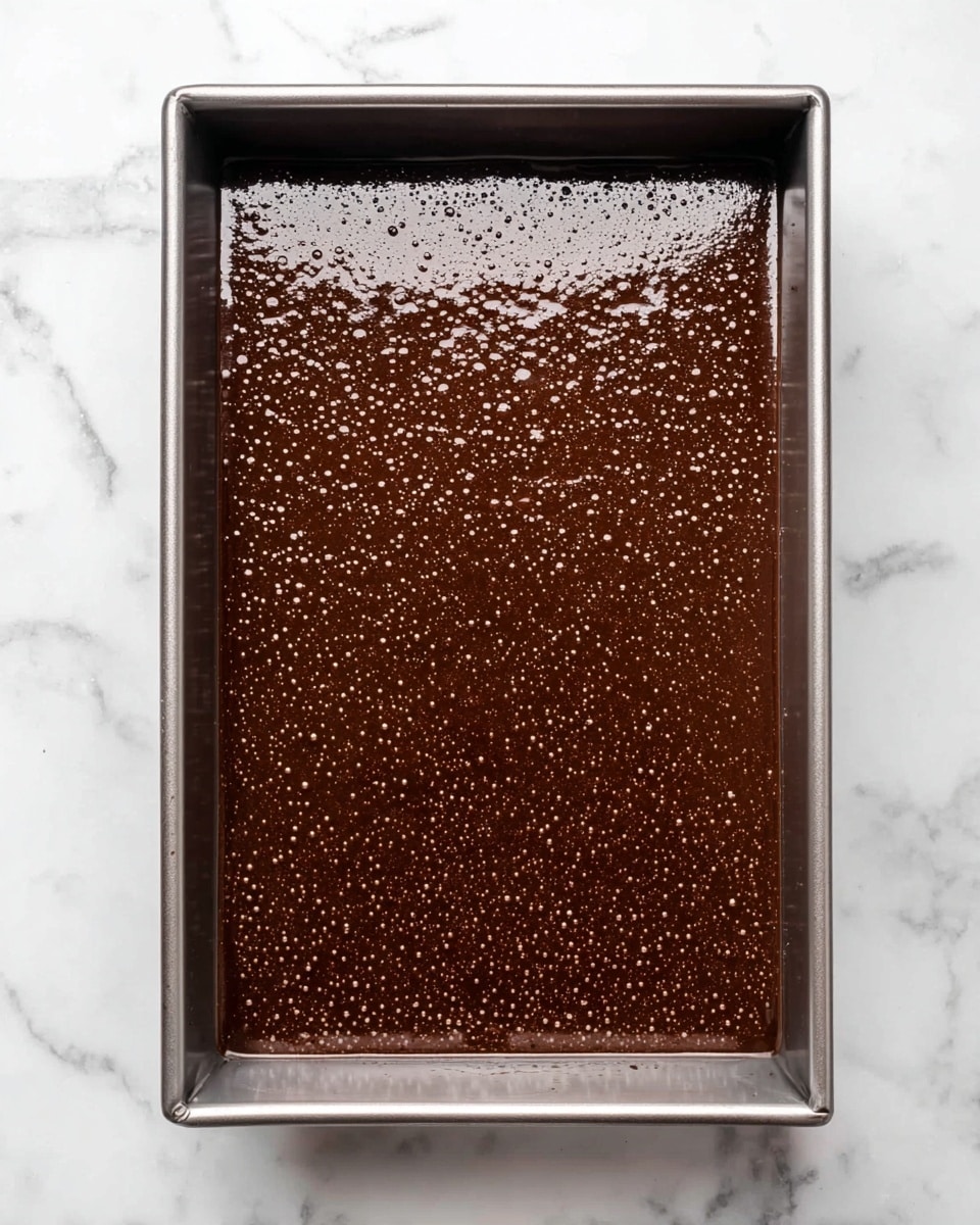 A metal rectangular baking pan filled with smooth, dark brown chocolate batter that has small bubbles all over the surface, and the batter is evenly spread to cover the entire pan. The pan sits on a white marbled surface. photo taken with an iphone --ar 4:5 --v 7