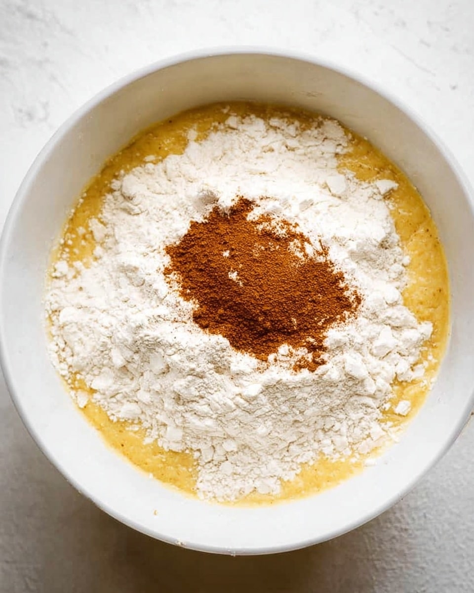 A white bowl contains three visible layers of ingredients: the bottom layer is a thick, smooth yellow batter partially visible around the edges; the middle layer is a generous mound of fine white flour evenly covering the batter; the top layer is a small round pile of brown cinnamon powder placed in the center of the flour. The bowl is set on a white marbled surface. Photo taken with an iphone --ar 4:5 --v 7