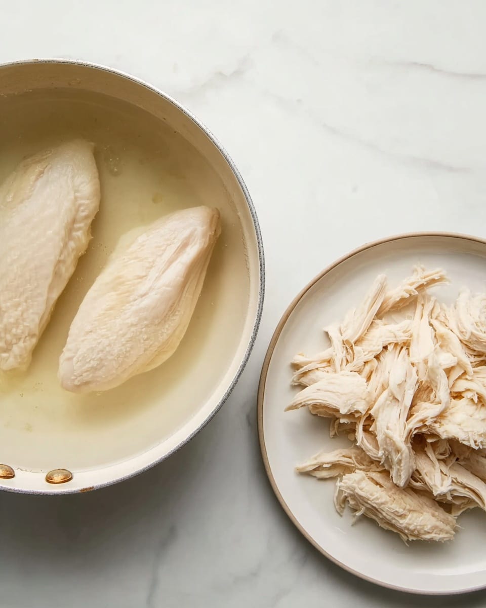 The image shows two light-colored chicken pieces simmering in a pan filled with clear water, placed on a white marbled surface. Next to the pan, on a plain white plate, there is shredded chicken arranged in loose strands with some bigger chunks on the right side of the plate. The colors are soft, mainly pale white and beige tones, with the smooth texture of the chicken breast and the slightly fibrous texture of the shredded chicken clearly visible. The scene is simple, clean, and focused on the cooking process, photo taken with an iphone --ar 4:5 --v 7