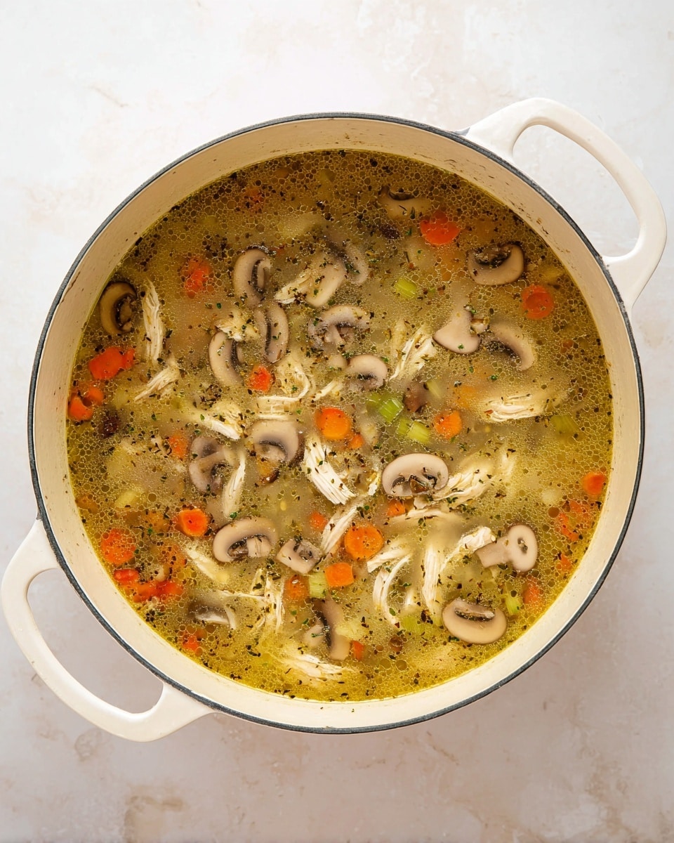 The image shows a large white pot filled with soup. The soup has a clear yellowish broth with floating pieces of white shredded chicken, sliced light brown mushrooms, small orange carrot cubes, and green celery slices. Some herbs sprinkled throughout the soup add fine specks of green. The pot sits on a white marbled surface, and the photo is taken from above, showing the round shape and handles on either side. photo taken with an iphone --ar 4:5 --v 7