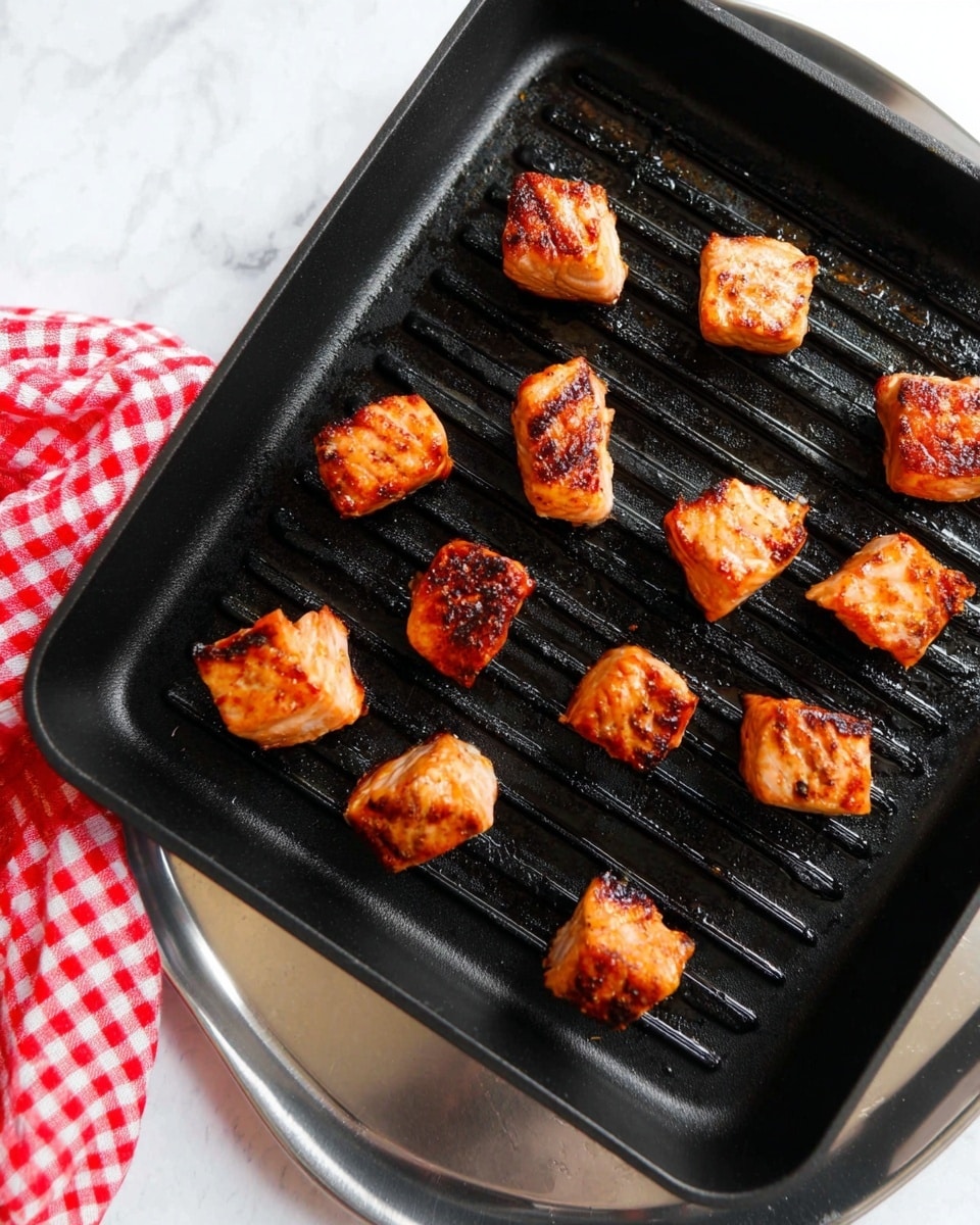 Several pieces of grilled salmon cubes are placed on a black grill pan with spaces between them, showing a golden-brown and slightly charred outer texture. The pan is resting on a shiny silver tray, sitting on a white marbled surface. To the left, a red and white checkered cloth is partially visible, adding a splash of color to the scene. The salmon pieces have an uneven, crispy look with some darker spots from grilling. photo taken with an iphone --ar 4:5 --v 7
