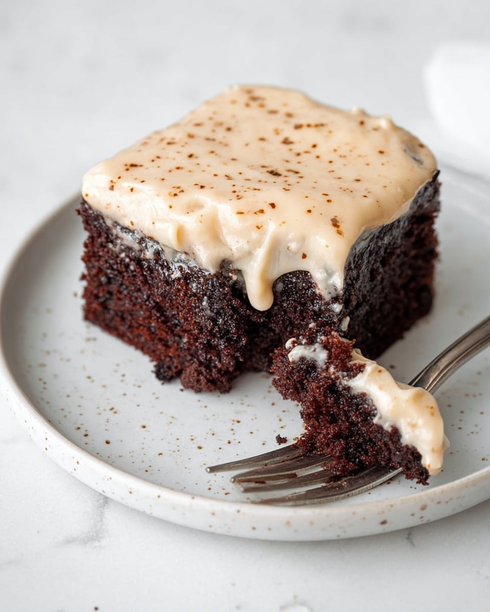 A single square piece of dark brown chocolate cake with a thick layer of light beige frosting on the top, slightly melting down the sides, sits on a white plate with small speckles. The frosting has small brown spots sprinkled over it. Next to the cake, a silver fork lies on the plate holding a bite-sized piece of cake topped with frosting. The background is a white marbled surface. photo taken with an iphone --ar 4:5 --v 7