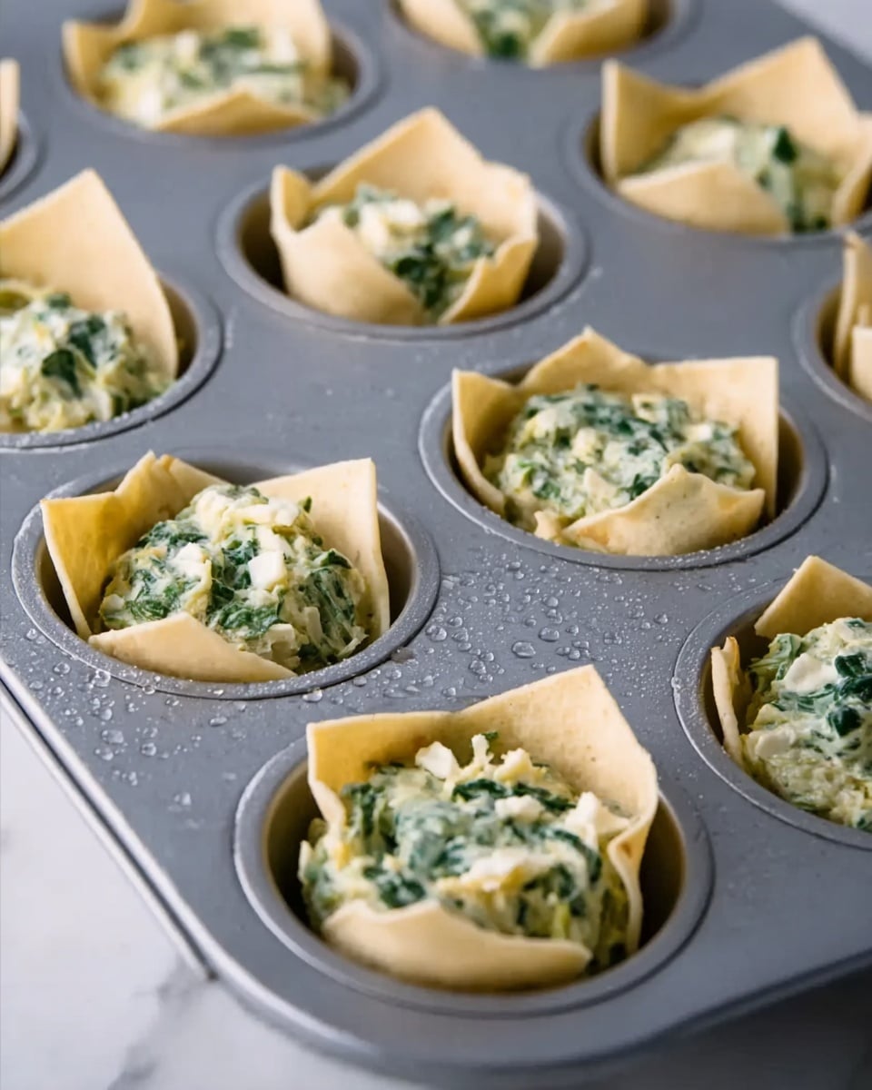 The image shows a close-up of a metal muffin tray filled with small, raw tortilla cups. Each cup has a light tan, thin tortilla layer forming a bowl shape. Inside each tortilla bowl, there is a thick, creamy mixture with visible bits of chopped green spinach and small chunks of pale cheese mixed in. The muffin tray sits on a white marbled surface with a few water droplets scattered around, giving a fresh and clean look. Photo taken with an iphone --ar 4:5 --v 7