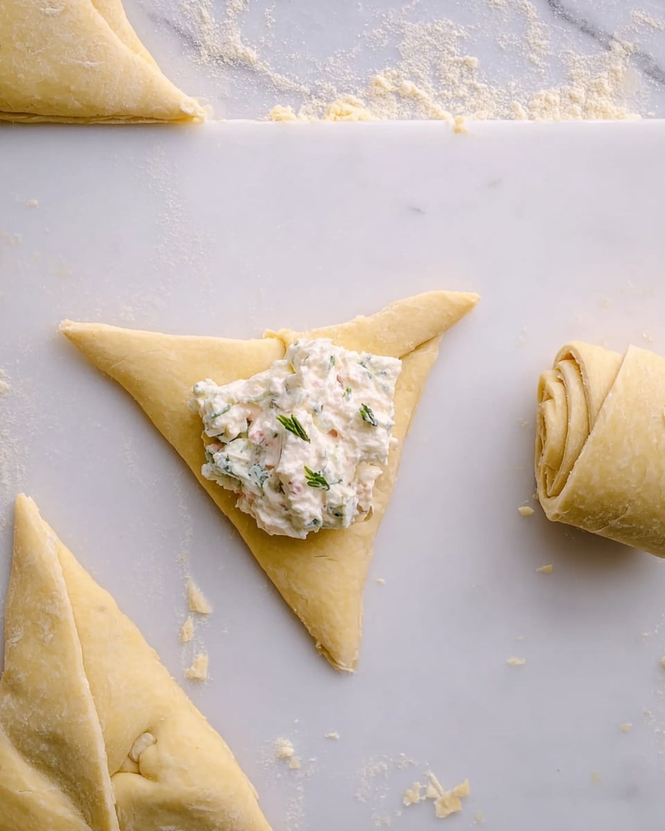 The image shows a white cutting board on a white marbled surface with pieces of dough shaped into triangles. One triangle dough piece is in the center with a dollop of creamy white filling mixed with small green herbs placed near its wider end. There is a rolled dough piece next to it on the right side, showing the start of a filled pastry. The dough has a light yellow color and a soft texture. Photo taken with an iphone --ar 4:5 --v 7