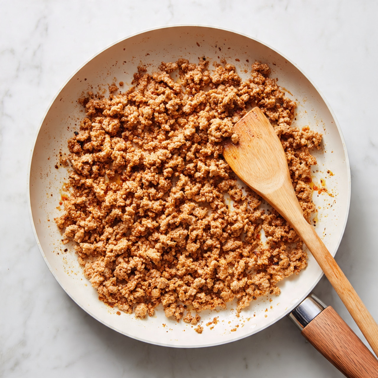 In a round white frying pan with a wooden handle, there is one layer of cooked ground meat that is light brown with some slightly darker bits around it, giving a textured and crumbly look. The pan is placed on a white marbled surface. A wooden spatula with some spices residue rests on the right side of the pan handle. The scene shows the meat in a single even layer, scattered across the pan bottom, with the meat looking dry and cooked evenly. Photo taken with an iphone --ar 4:5 --v 7