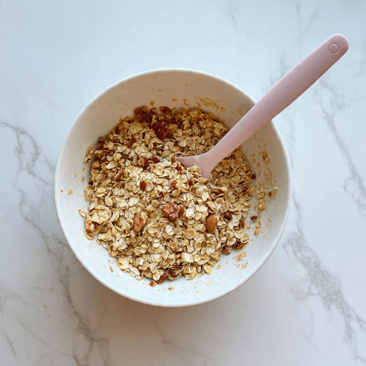 A white bowl filled with a mix of light brown oats and golden brown nuts, with small darker brown chunks scattered throughout. A pale pink spatula rests inside the bowl on the right side, partially covered by the oats mixture. The bowl sits on a white marbled surface, and the lighting highlights the texture of the oats and nuts clearly. photo taken with an iphone --ar 4:5 --v 7