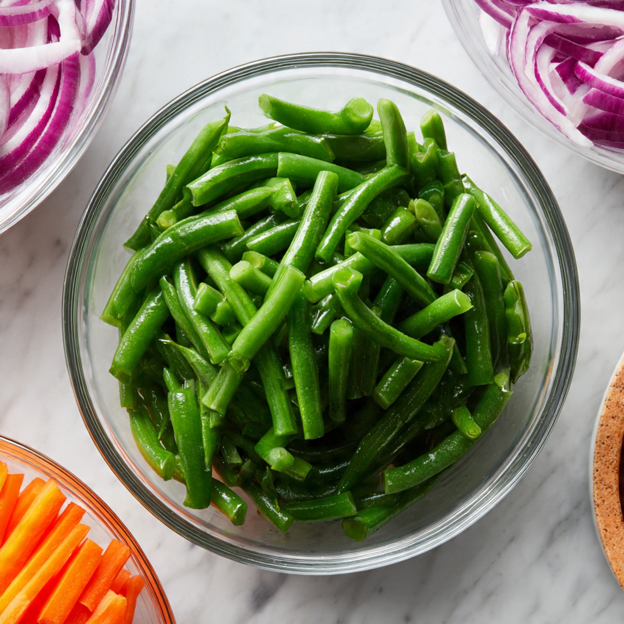 A clear glass bowl filled with bright green fresh green beans, trimmed and cut into pieces, sitting on a white marbled surface. In the top left corner, there is a partial view of another clear glass bowl filled with sliced red onions showing purple and white layers, and at the bottom left corner there is a glimpse of a clear glass bowl containing vibrant orange carrot sticks. The image is bright and crisp, showing the fresh texture and color of the vegetables, photo taken with an iphone --ar 4:5 --v 7