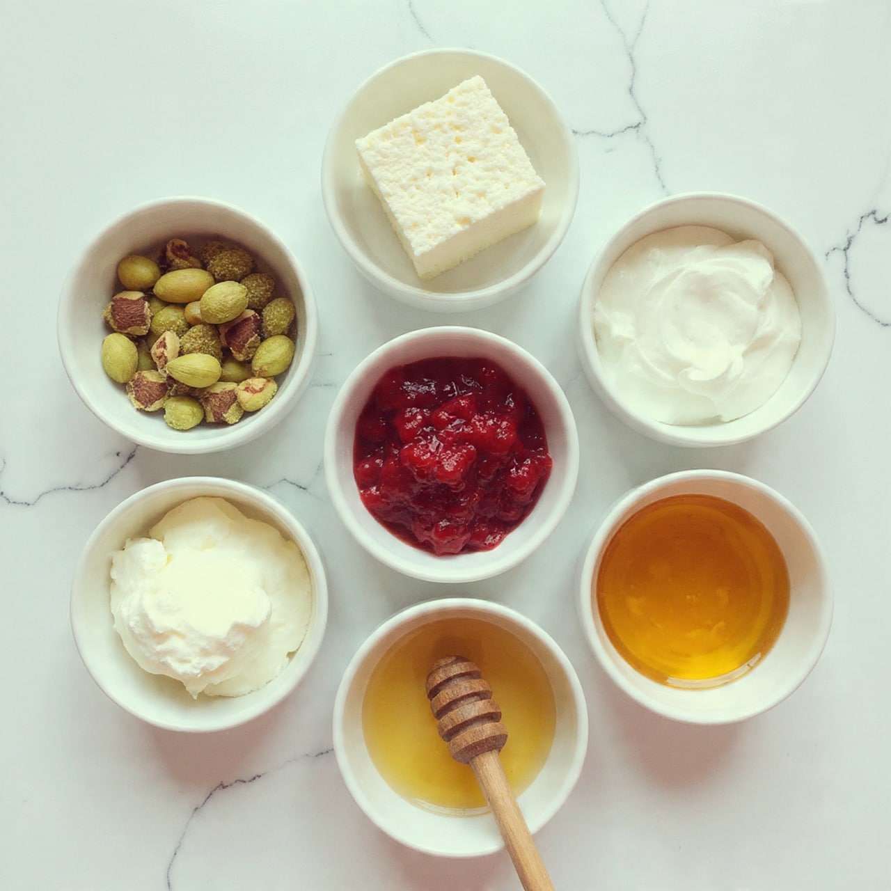 The image shows five small white bowls on a white marbled surface, each filled with a different ingredient. From top to bottom and left to right: one bowl contains green and brown pistachios with a rough texture; next to it is a bowl with a full round block of white feta cheese in a clear liquid; below the pistachios is a bowl filled with bright red, chunky cranberry sauce; next to the cranberries is a bowl with a square piece of smooth, creamy white cream cheese; at the bottom left, there is a bowl filled with golden honey, glossy and thick with a wooden honey dipper resting inside. The bowls are arranged in a loose cluster. photo taken with an iphone --ar 4:5 --v 7