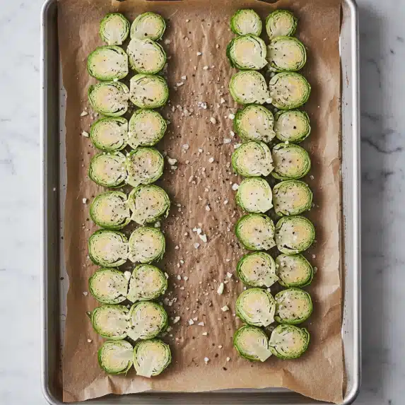 The image shows sliced Brussels sprouts, about two layers thick, placed on a baking sheet lined with brown parchment paper. The Brussels sprouts are bright green on the outer edges with a pale creamy center, coated with small bits of minced garlic and sprinkled with coarse salt and black pepper. The baking sheet is silver and shows signs of use. The setup is on a white marbled surface. photo taken with an iphone --ar 4:5 --v 7