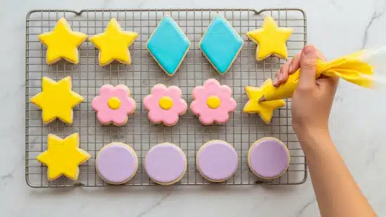 The Best Rolled Sugar Cookies Recipe 5 The image shows a white marbled surface with a metal cooling rack holding twelve sugar cookies in three different shapes: stars, diamonds, flowers, and circles. The star-shaped cookies have smooth, bright yellow icing, while the diamond-shaped cookies are covered in a shiny bright blue icing. The flower-shaped cookies have soft pink icing with a small yellow circle in the center, and the round cookies are coated evenly with a pastel purple icing. On the right side of the rack, a woman’s hand is squeezing bright yellow icing from a piping bag to decorate an undecorated star-shaped cookie. The lighting is soft, and the overall scene looks clean and colorful photo taken with an iphone --ar 4:5 --v 7