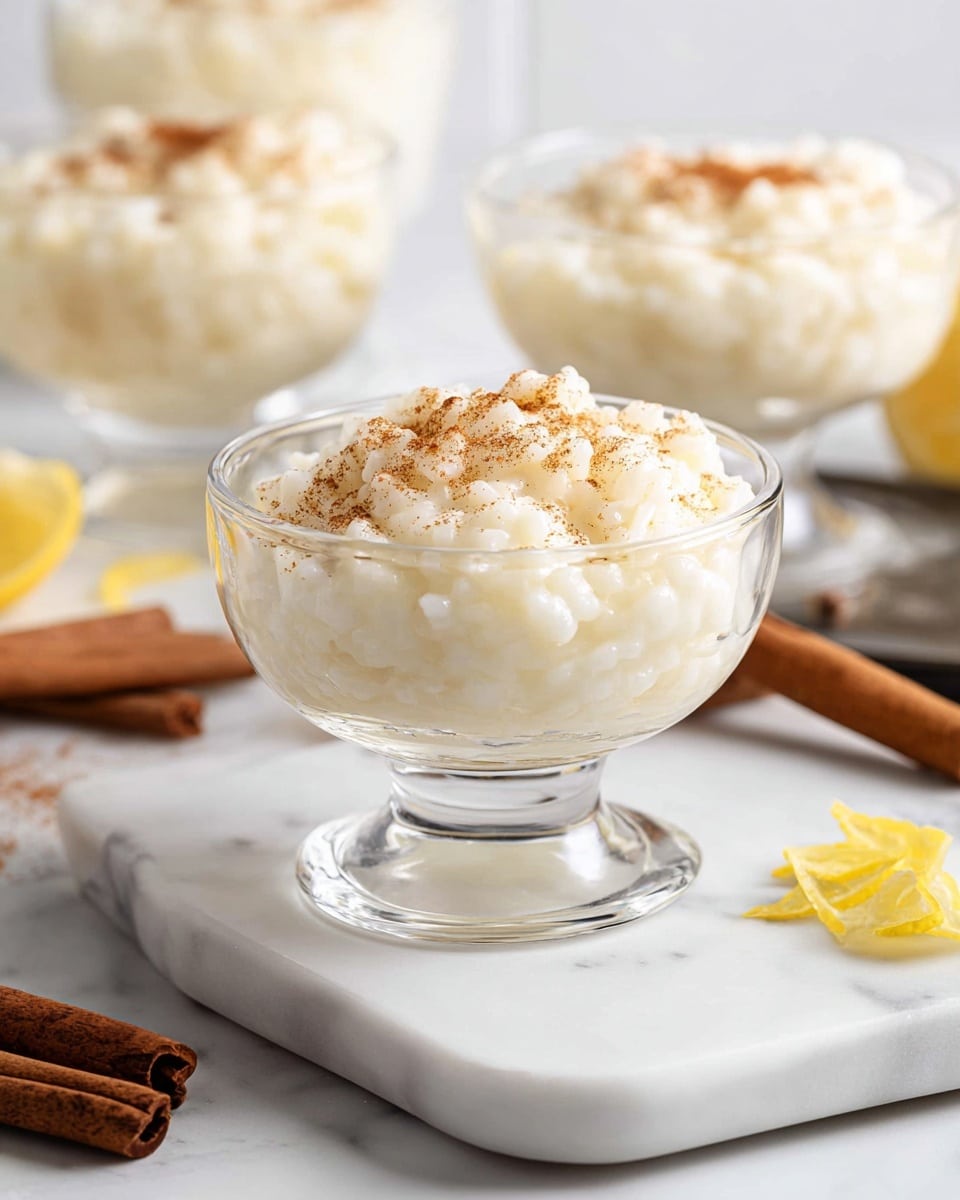 The image shows clear glass dessert cups filled with a creamy, white rice pudding that has a soft and slightly lumpy texture. Each cup holds one layer of the rice pudding topped with a light sprinkling of brown cinnamon powder. The cups are placed on a white marbled surface with two cinnamon sticks and a small twist of yellow lemon peel lying next to one of the cups. The background features a soft white tiled wall, adding to the clean and bright look. photo taken with an iphone --ar 4:5 --v 7