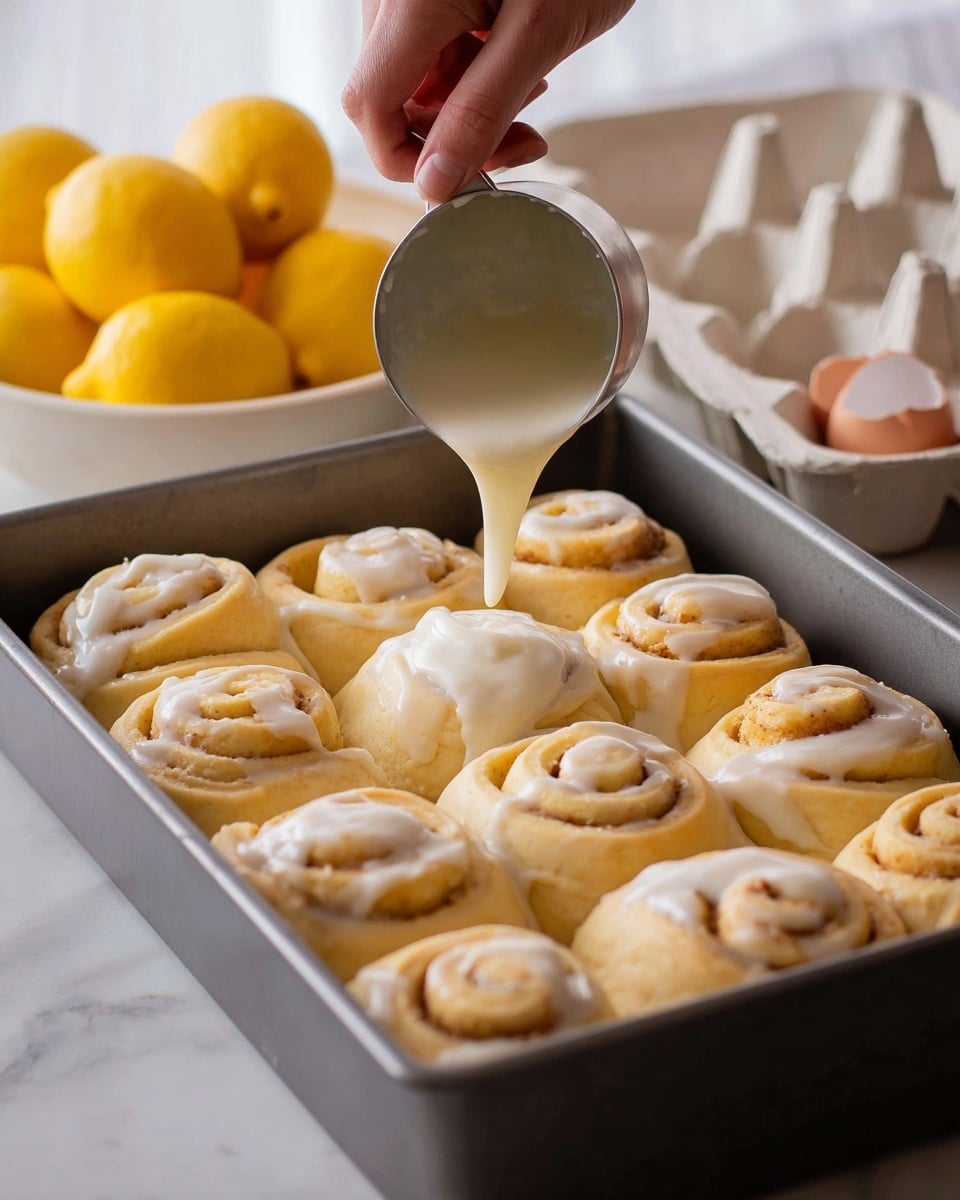 A baking pan is filled with twelve soft, light beige dough rolls arranged in three rows and four columns. A woman's hand is pouring smooth, white icing from a small metal measuring cup over the rolls, creating a glossy layer that flows and spreads over their circular tops and sides. The rolls show distinct spiral layers of dough that are slightly puffy and textured. In the background, there is a white bowl filled with bright yellow lemons and a white egg carton with some brown eggshells visible, all placed on a white marbled surface. Photo taken with an iphone --ar 4:5 --v 7