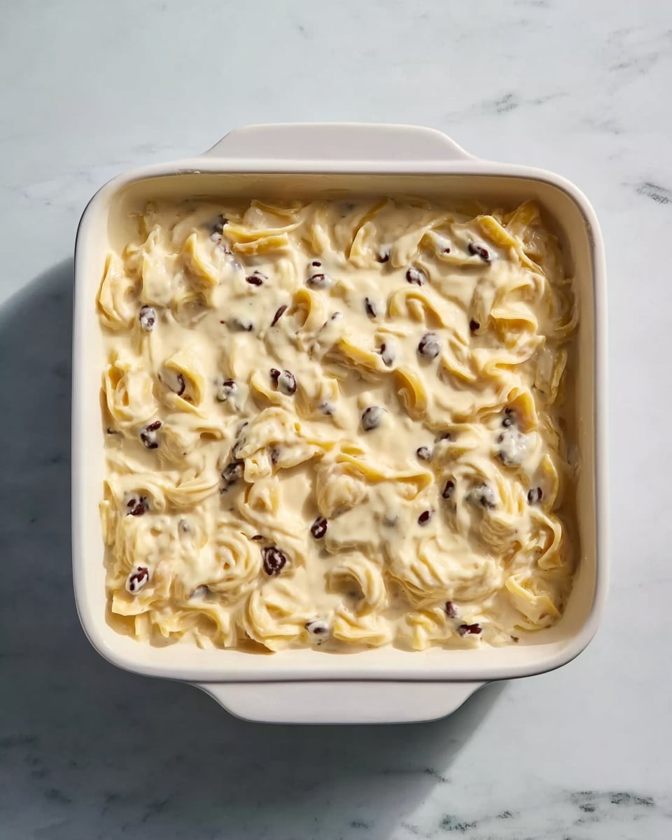 A square white ceramic dish filled with a creamy mixture of pale yellow pasta swirls and small dark beans evenly spread inside. The sauce is thick and covers the pasta fully, creating a smooth, slightly lumpy texture on top. The dish is placed on a white marbled surface, showing some soft light reflections on the sauce and subtle shadows from the dish edges. Photo taken with an iphone --ar 4:5 --v 7