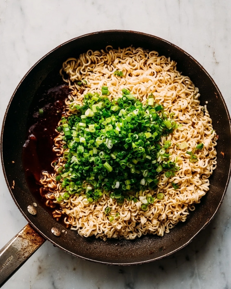 A close-up of a dark, round frying pan on a white marbled surface filled with instant noodles. The noodles are pale yellow, curly, and piled mostly on the right side of the pan. On the left side of the pan, there is a dark brown sauce pooling underneath the noodles. On top of the noodles and sauce, in the center, is a mound of chopped green onions that are fresh and bright green. The pan handle extends horizontally to the left. photo taken with an iphone --ar 4:5 --v 7