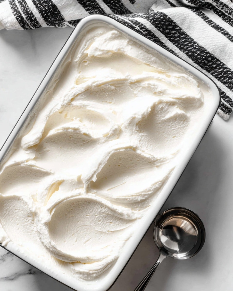 A close-up image shows a rectangular white baking dish filled with a thick, creamy white mixture that has smooth swirls and soft peaks on the surface, giving it a light, fluffy texture. The dish is placed on a white marbled surface, with a striped black and white cloth partially visible in the upper right corner. A shiny metal scoop with a round edge rests on the right side of the dish. photo taken with an iphone --ar 4:5 --v 7