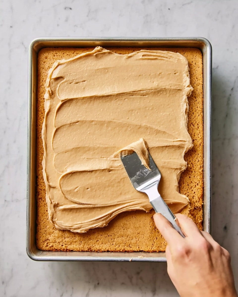 The image shows a rectangular baked cake layer with a golden brown color inside a metal tray. A woman's hand is holding a metal spatula spreading a thick, smooth layer of light tan frosting evenly over the cake's surface. The texture of the frosting looks creamy and consistent, covering the top fully but not the sides yet. The tray is placed on a white marbled surface. Photo taken with an iphone --ar 4:5 --v 7