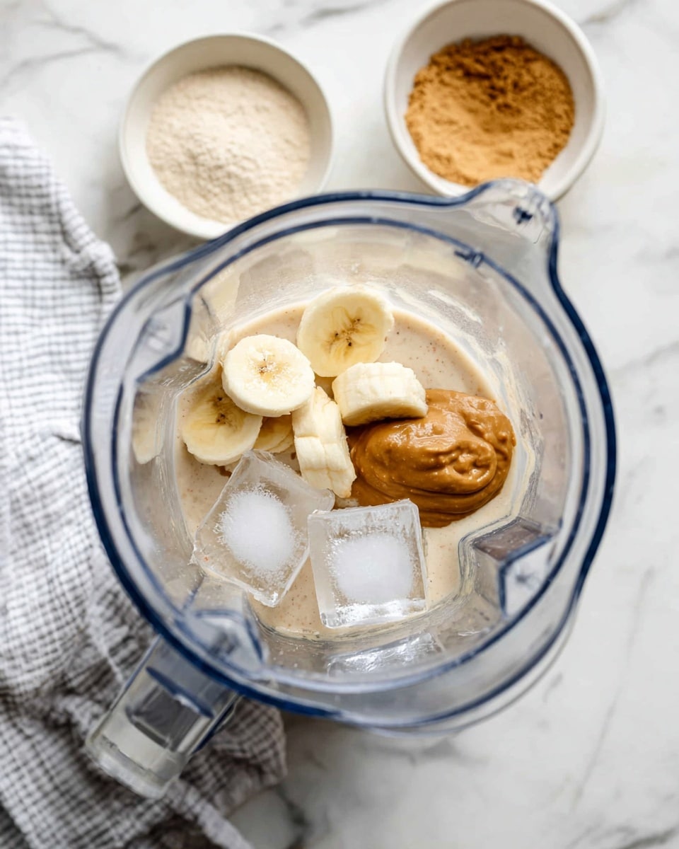 A clear blender jar is shown from above, filled with several ingredients in distinct layers: at the bottom are creamy light beige liquid and powder, topped with light yellow banana slices, a dollop of brown nut butter, and two large transparent ice cubes. Around the blender, there are two small white bowls, one with pale beige powder and another with light brown nut butter sauce. A white and gray striped cloth sits near the blender base on a white marbled surface. Photo taken with an iphone --ar 4:5 --v 7
