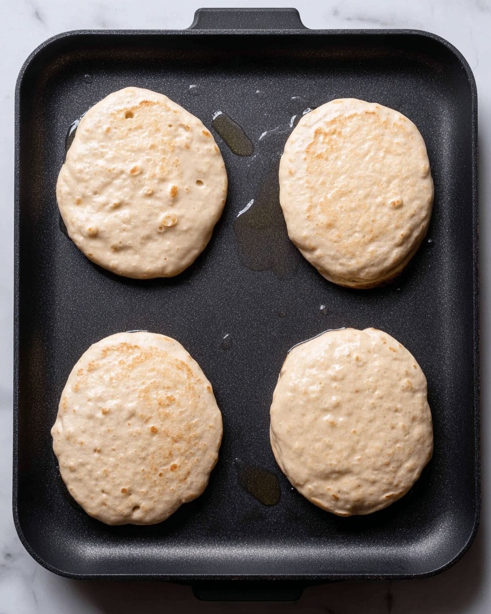 The image shows four small pancakes cooking on a dark non-stick griddle pan, arranged in a square pattern. Each pancake is thick and round, with a light beige color and visible small bubbles on the surface, indicating they are in the early stage of cooking. The pancakes have a slightly uneven texture with some small lumps. The griddle pan has droplets of oil or water around the pancakes, and the background features a white marbled surface. photo taken with an iphone --ar 4:5 --v 7