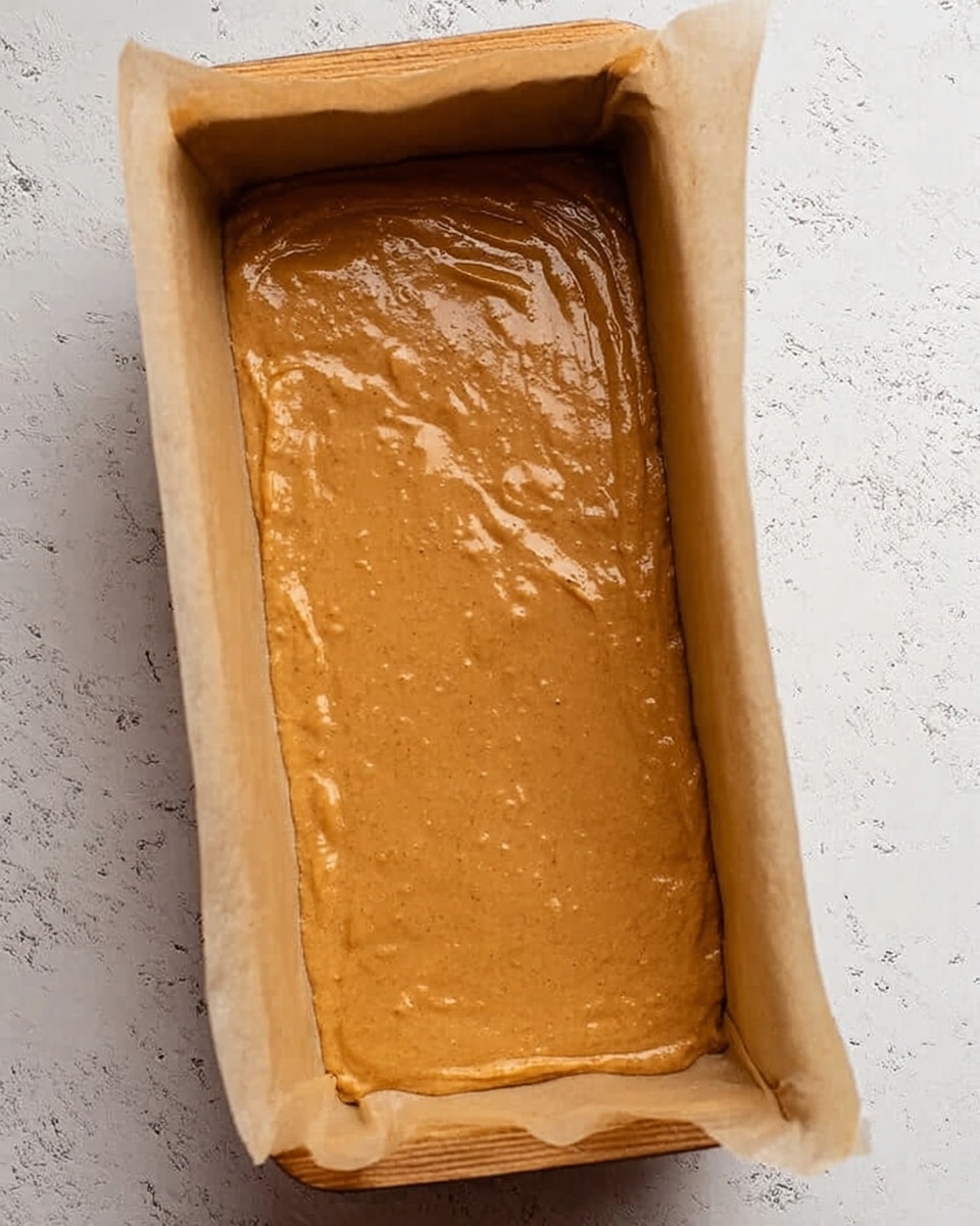 A rectangular baking tray lined with light brown parchment paper holds smooth, light brown batter spread evenly inside. The batter’s texture is creamy with some small lumps, filling the tray almost to the top edges. The tray is placed on a white marbled surface that adds a clean background to the image. photo taken with an iphone --ar 4:5 --v 7