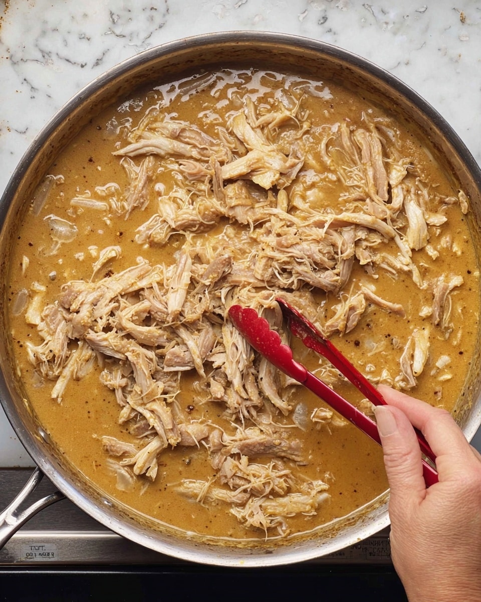 A close-up view of a silver pan filled with a thick, light brown sauce. Inside the sauce are shredded pieces of light beige meat with some browned edges, mixed evenly throughout the sauce in a single layer. A woman's hand is holding red-tipped tongs, lifting and stirring the meat in the sauce. The pan is placed on a stove, and the surface beneath has a white marbled texture. Photo taken with an iphone --ar 4:5 --v 7