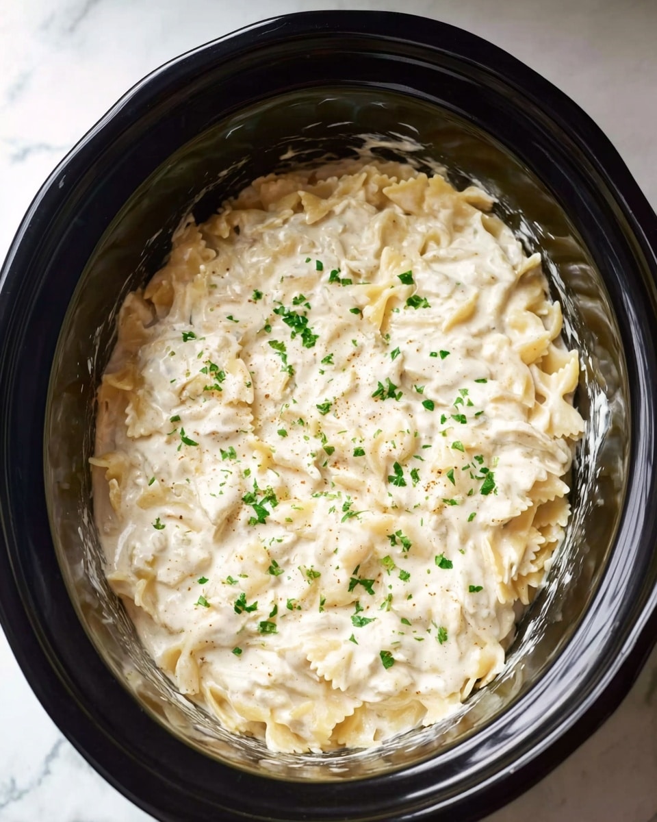 The image shows a close-up top view of a slow cooker filled with creamy white sauce pasta. The dish has one layer of bow-tie pasta mixed evenly with a thick, smooth white sauce, sprinkled lightly with small green herb pieces for color contrast. The inside of the slow cooker is black, and the creamy pasta almost fills the container, with little sauce visible on the sides. The background is a white marbled surface. Photo taken with an iphone --ar 4:5 --v 7