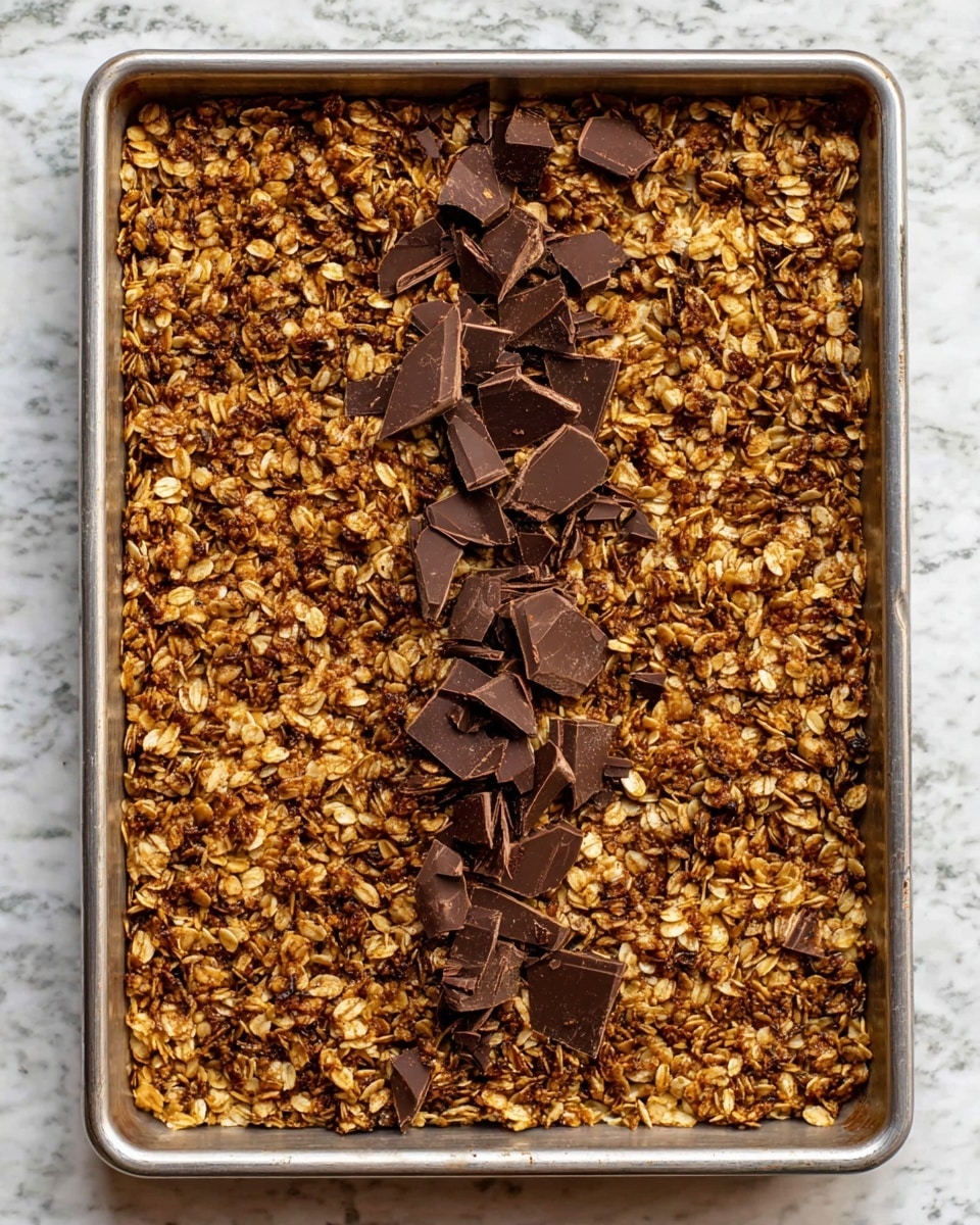 A large rectangular metal baking tray filled with a single thick layer of baked granola that is golden brown with visible oats and puffed grains throughout. On top, there is a row of roughly chopped dark chocolate pieces scattered across the middle, adding a dark, rich contrast to the textured granola base around it. The tray is placed on a white marbled surface, showing some slight baked spots near the edges. The granola has a crisp, crumbly look and the chocolate pieces vary in size with sharp edges, partially melting into the warm granola beneath photo taken with an iphone --ar 4:5 --v 7