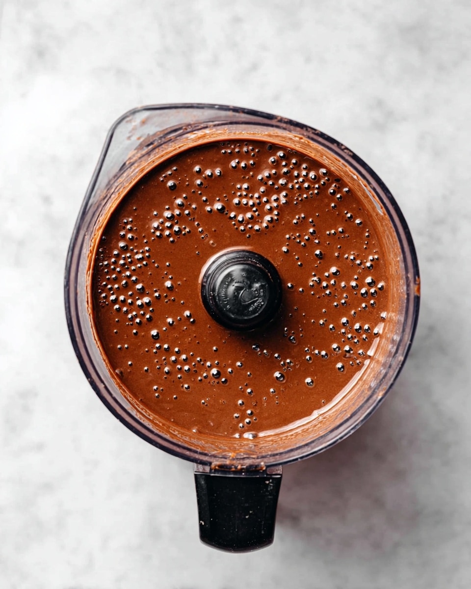 The image shows a top view of a blender container filled with a thick, smooth chocolate mixture. The mixture is dark brown with small bubbles evenly spread across the surface, giving it a rich and creamy texture. The blender container is clear with a black lid and handle, placed on a white marbled surface. The scene is well-lit, highlighting the shine and thickness of the chocolate mix. photo taken with an iphone --ar 4:5 --v 7