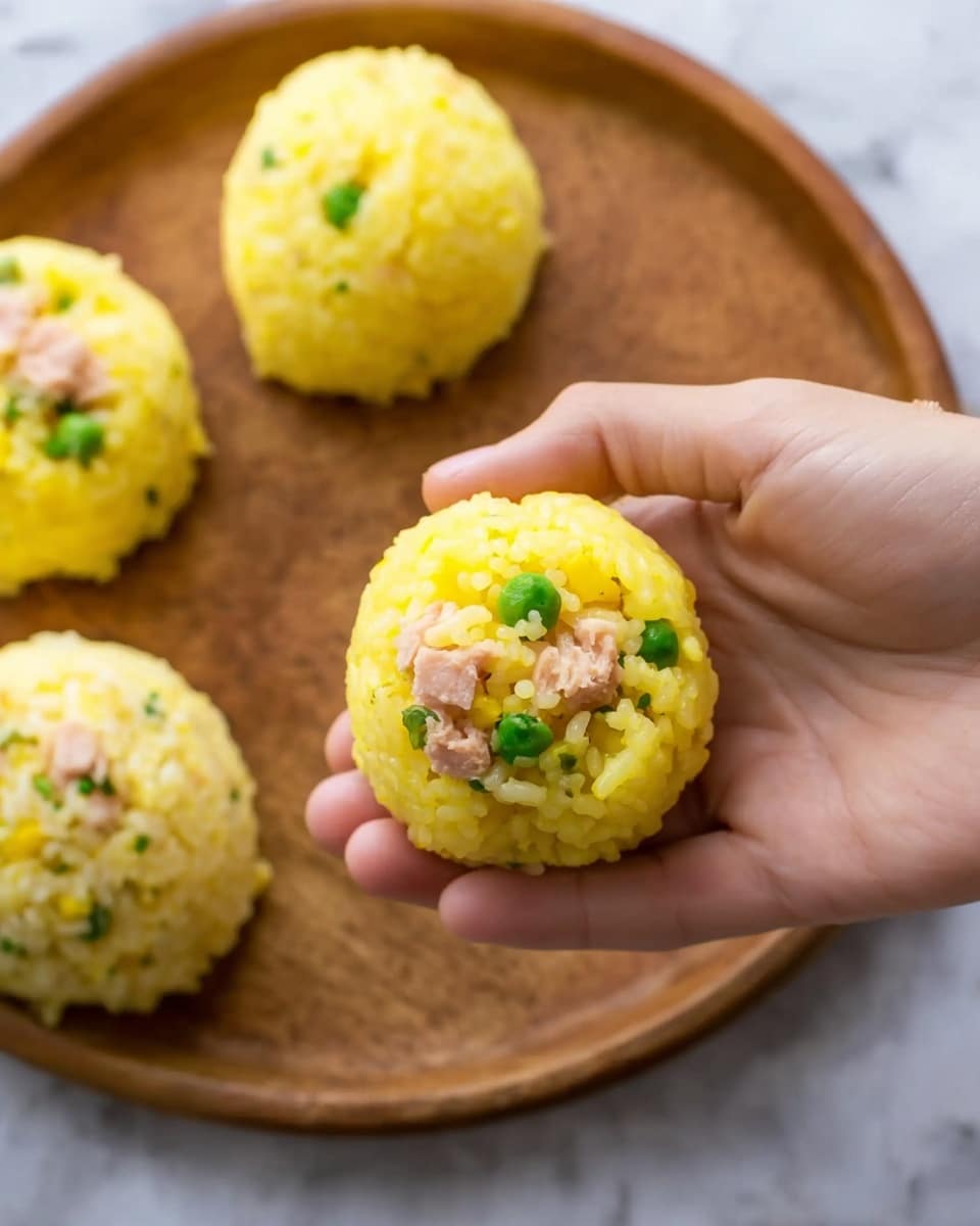 The image shows a close-up of a woman's hand holding a ball of yellow rice on top of a wooden plate. The rice ball is round and the rice grains are clearly visible with a soft texture. Inside the rice ball, there are green peas and small pieces of light pink tuna, adding color contrast to the yellow rice. In the background, more rice balls with similar fillings rest on the wooden plate. The whole scene is placed on a white marbled surface. photo taken with an iphone --ar 4:5 --v 7