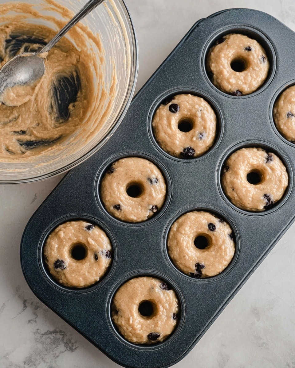 A close-up of a dark baking tray filled with six light brown batter portions for small ring-shaped cakes or donuts, each with small dark spots that look like blueberries, placed in individual round molds with a cylindrical center. To the left, a clear glass mixing bowl contains more of the same batter with dark spots, with a silver spoon partially visible inside. The tray and bowl are set on a white marbled surface. photo taken with an iphone --ar 4:5 --v 7