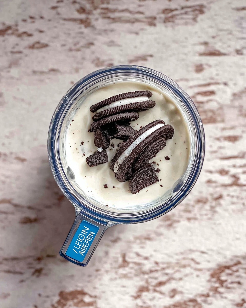 A top view of a clear blender jar filled with a thick white creamy mixture, topped with several pieces of dark brown and white sandwich cookies placed in the center, sitting on a white marbled surface. The jar has a blue handle with white text reading