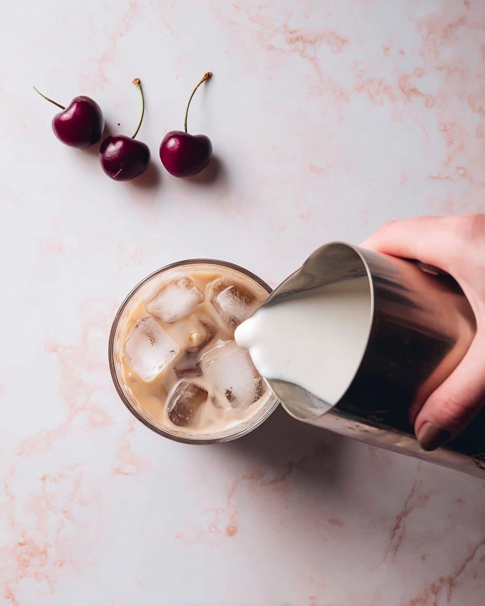 A clear cup filled with three floating ice cubes and light brown coffee is shown from above on a white marbled texture. A woman's hand holds a metal pitcher pouring creamy foamy milk into the cup from the right side. Two dark red cherries with green stems lie in the upper left corner on the white marbled surface. photo taken with an iphone --ar 4:5 --v 7