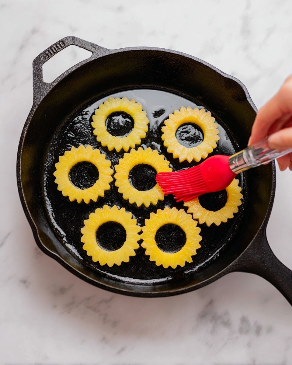 Seven yellow, scalloped-edged rings with hollow centers are arranged in a single layer in a black cast iron skillet. The skillet has some shiny oil visible beneath the rings, creating a reflective surface. A woman's hand is using a red silicone brush with a clear handle to spread oil or glaze over the center ring. The skillet sits on a white marbled surface, and the overall image shows a close top-down view. photo taken with an iphone --ar 4:5 --v 7