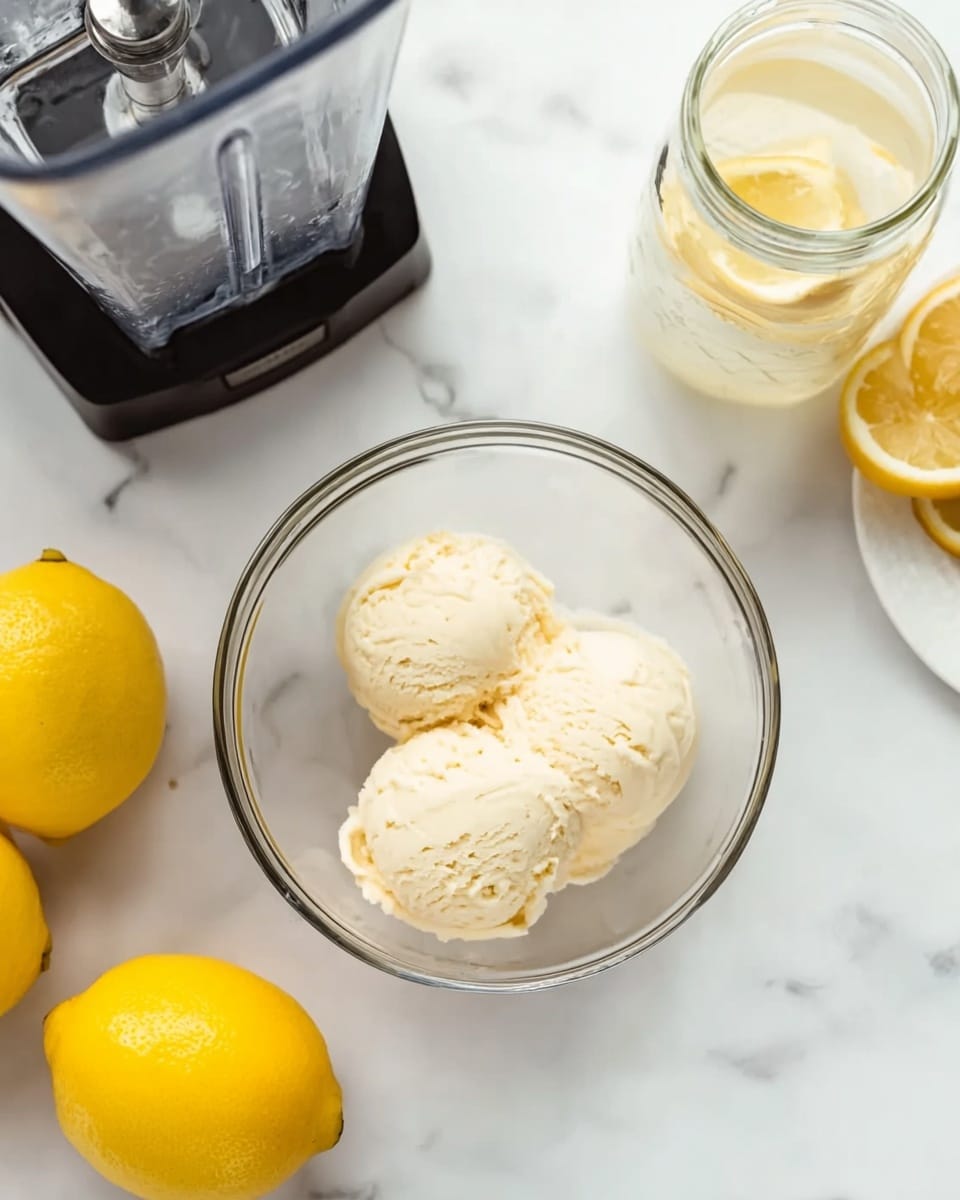 A clear glass bowl in the center holds three scoops of pale yellow ice cream with a creamy texture, sitting on a white marbled surface. To the left, there are four bright yellow lemons with smooth skin, while on the right side, a glass jar filled with a whitish-yellow liquid is placed next to partially visible lemon slices. Above the bowl, part of a blender with a transparent container and black base is shown. The overall image is bright and clean, with simple, fresh ingredients clearly visible, photo taken with an iphone --ar 4:5 --v 7