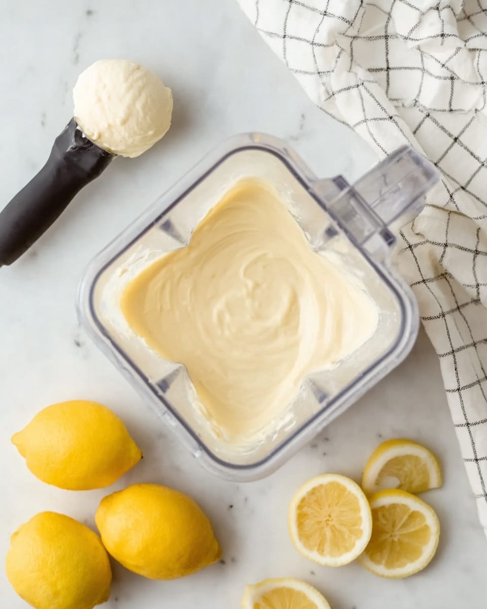 The image shows a clear blender container filled with a smooth, thick, pale yellow creamy mixture evenly spread inside. Below the blender, there are three whole bright yellow lemons placed on a white marbled surface, and half a lemon with slices arranged on the right side. Near the top left, a black ice cream scooper holds a round scoop of light cream-colored mixture. A white cloth with a gray grid pattern is spread in the upper right corner on the white marbled background. The overall scene looks clean and bright. photo taken with an iphone --ar 4:5 --v 7