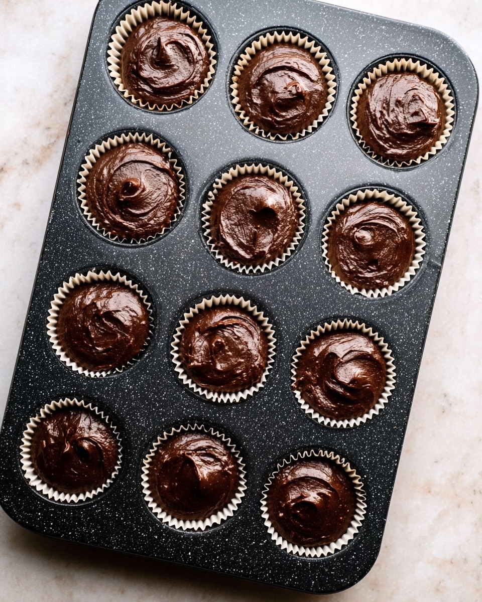 A black speckled muffin tray filled with twelve white paper liners, each filled with a smooth, dark chocolate batter that has a thick and swirled texture, almost full to the top of the liners. The tray is set on a white marbled surface. The batter is rich and shiny, with slight peaks and swirls in each muffin cup. photo taken with an iphone --ar 4:5 --v 7