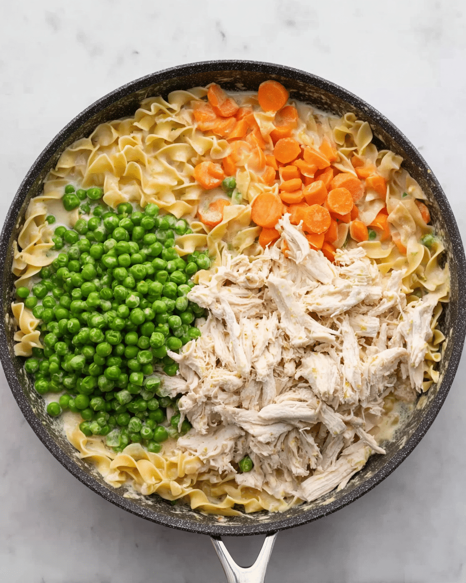 The image shows a dark round pan on a white marbled surface filled with four main layers of ingredients not yet mixed. On the bottom layer, there are wide egg noodles mixed with cooked, soft orange carrot slices in a light cream-colored liquid. On the top right, there is a large pile of shredded pale cooked chicken. On the top left, bright green frozen peas form a thick pile. The pan handle extends downward and is light silver in color. Photo taken with an iphone --ar 4:5 --v 7